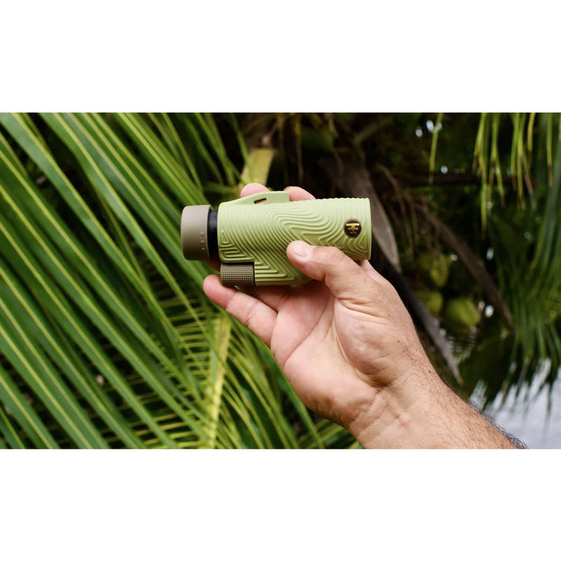 Load image into Gallery viewer, A hand holding a compact, olive green monocular with a textured grip and a small gold shield logo, set against a background of green palm leaves.